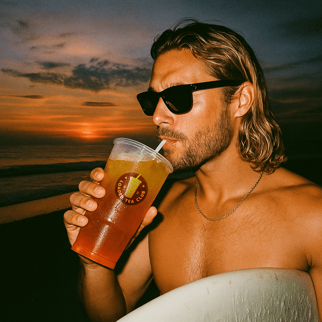 Man enjoying a loaded tea beverage at sunset on the beach, wearing sunglasses and holding a surfboard.