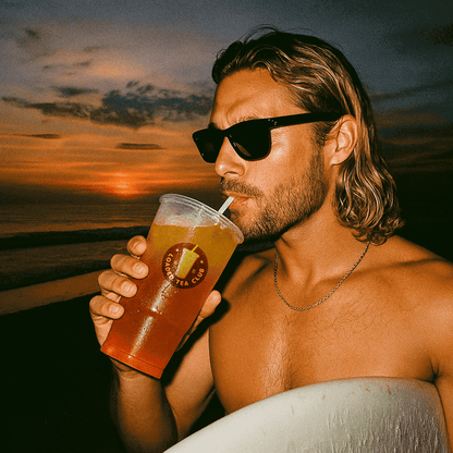 Man enjoying a loaded tea drink at sunset, wearing sunglasses and holding a surfboard.